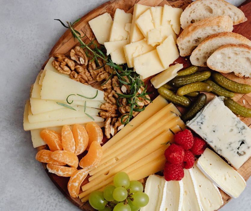 Charcuterie board with various hard and soft cheeses, walnuts, grapes, raspberries, oranges, pickles, and bread slices.