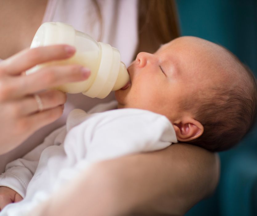A newborn baby with eyes closed being fed from a plastic bottle held by an adult.