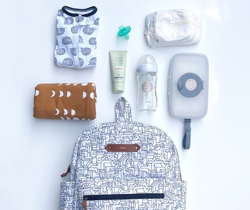 Daycare essentials including a backpack, pajamas, diapers, wipes, a bottle, cream, and a pacifier on a white surface.