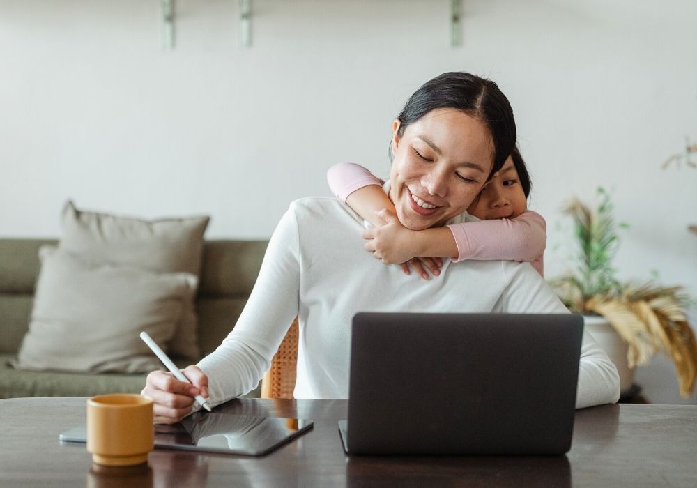 A woman works on a laptop and tablet at home while a young child hugs her from behind.