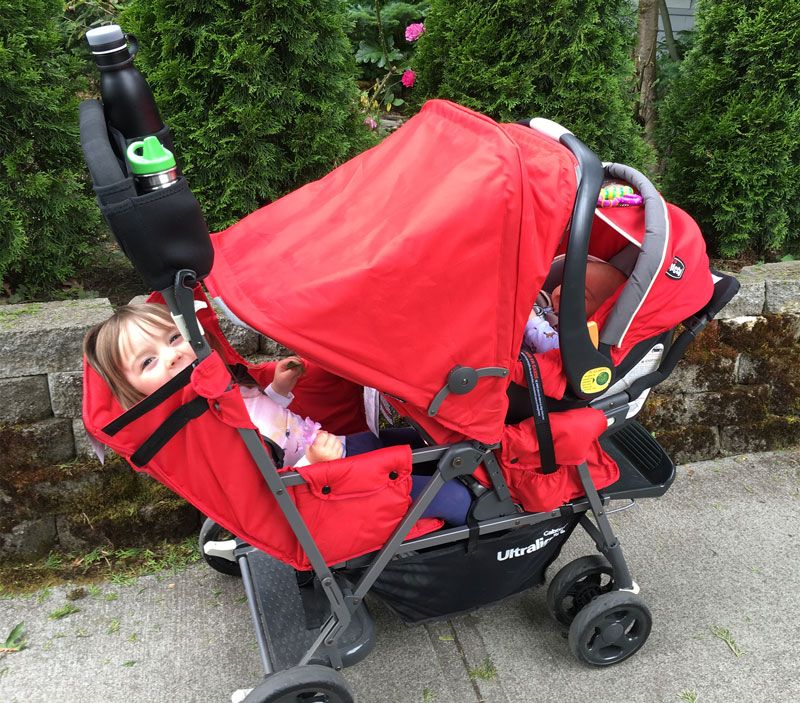 Red Joovy Caboose double stroller with an infant car seat in front and a toddler sitting in the rear seat.