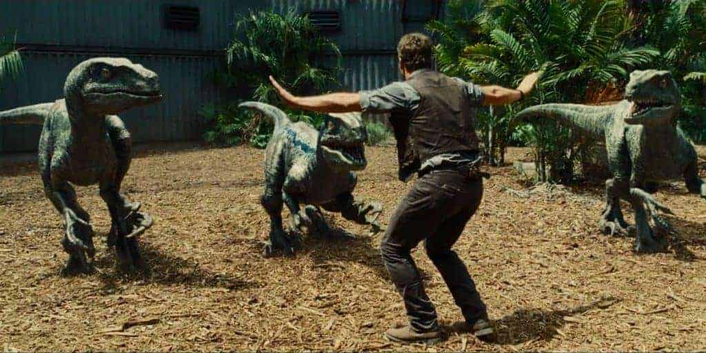 Owen Grady stands with arms outstretched, taming three velociraptors in an enclosure.