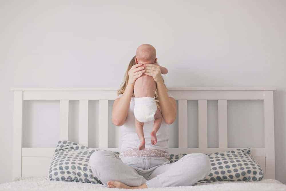 Woman sitting cross-legged on a bed, holding a baby in a diaper up in front of her face.