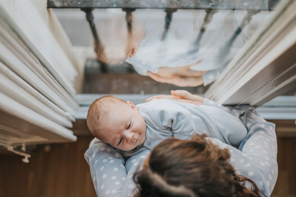 Top-down view of a mother holding her newborn baby near a window, with their reflection visible in the glass.