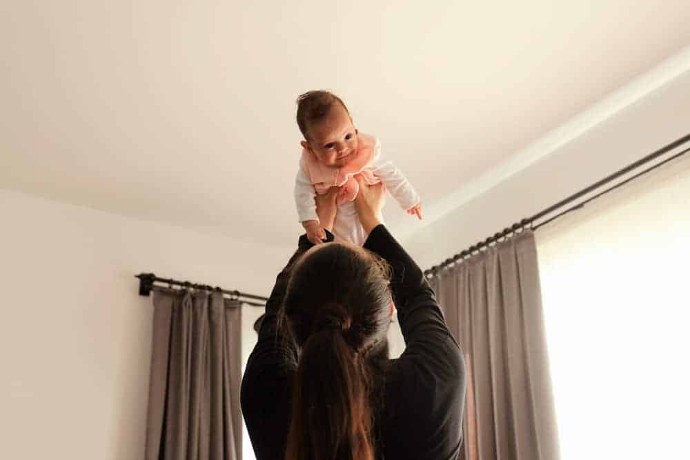 A mother in a black shirt lifts her baby in the air toward the ceiling in a bright room.