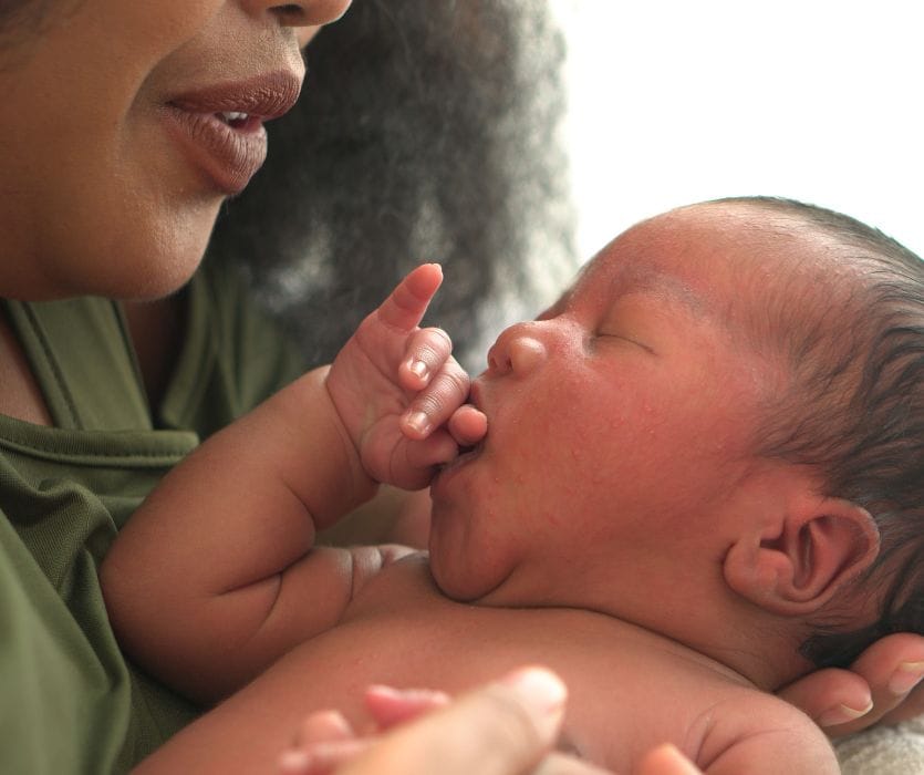 A close-up of a mother holding her sleeping newborn baby who has their thumb in their mouth.