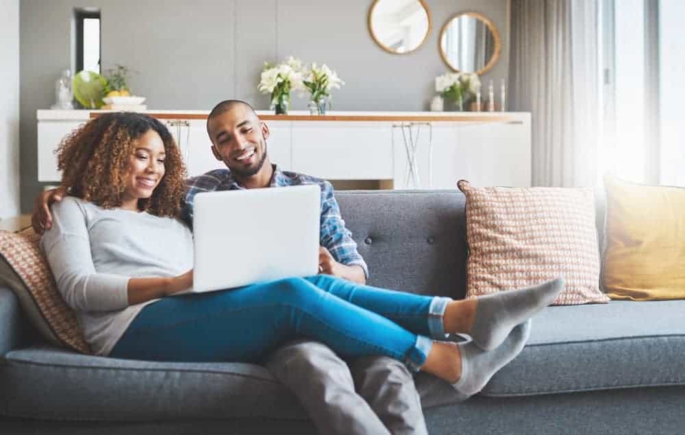 A smiling man and woman sit together on a gray sofa looking at a laptop computer.