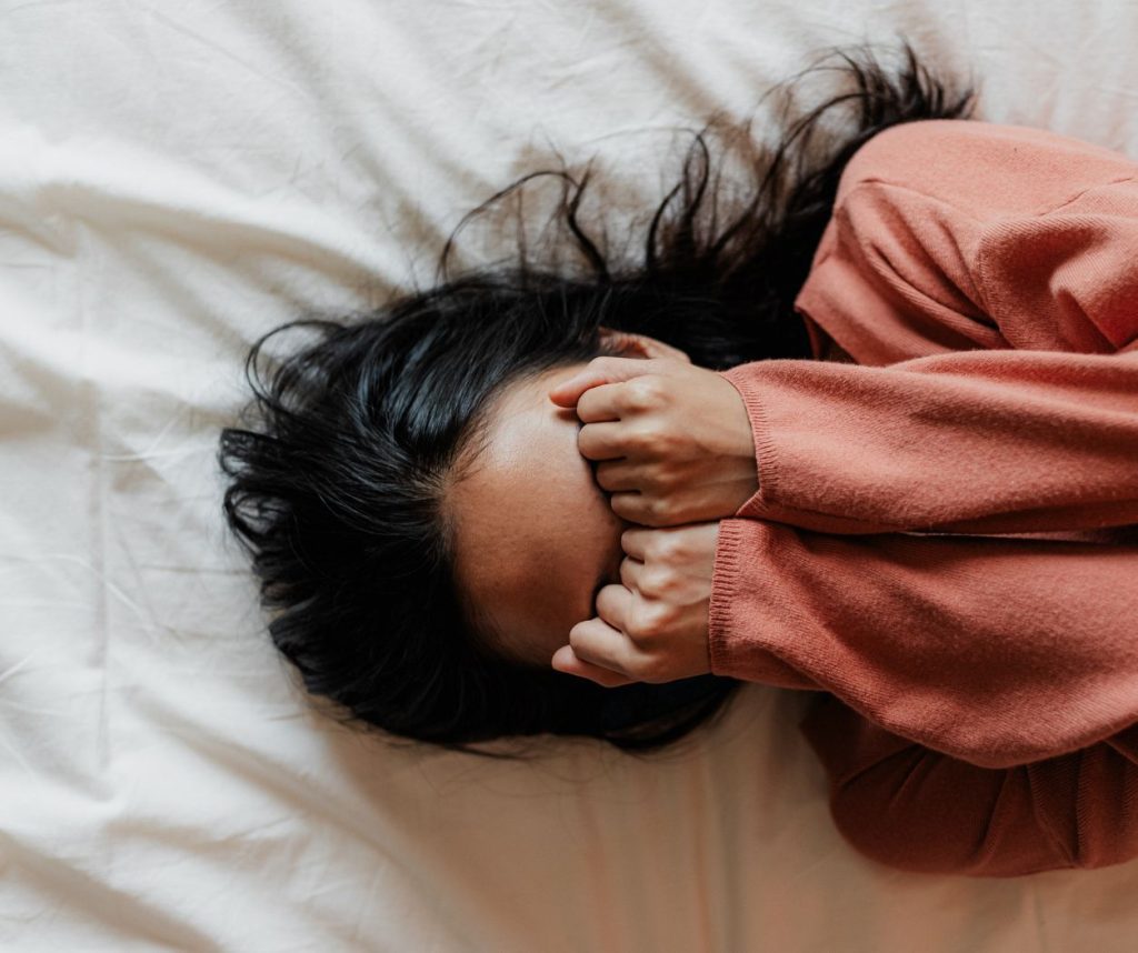 Overhead view of a person lying on white sheets covering their eyes with their hands.