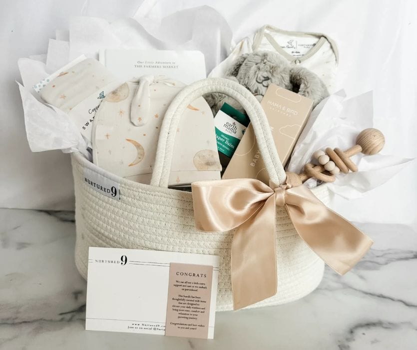 Baby shower gift basket with a plush bunny, wooden rattle, onesie, and skincare products in a cream rope basket with a bow.