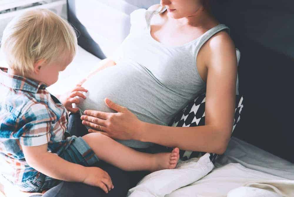 A toddler touches the belly of a pregnant woman wearing a grey tank top as they sit on a bed.