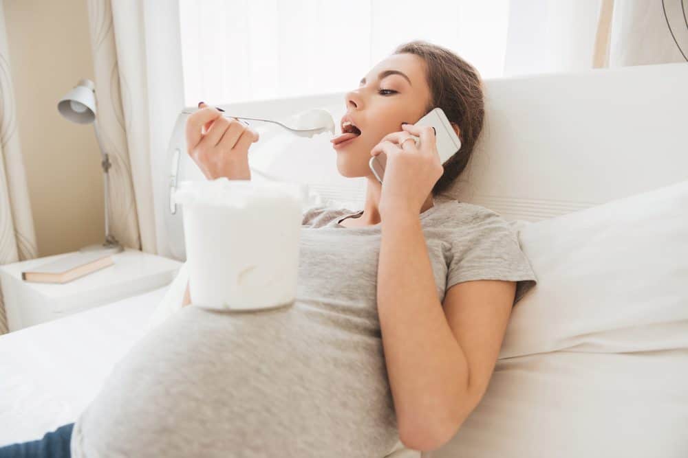 Pregnant woman reclining in bed, eating ice cream from a tub on her belly while talking on a smartphone.