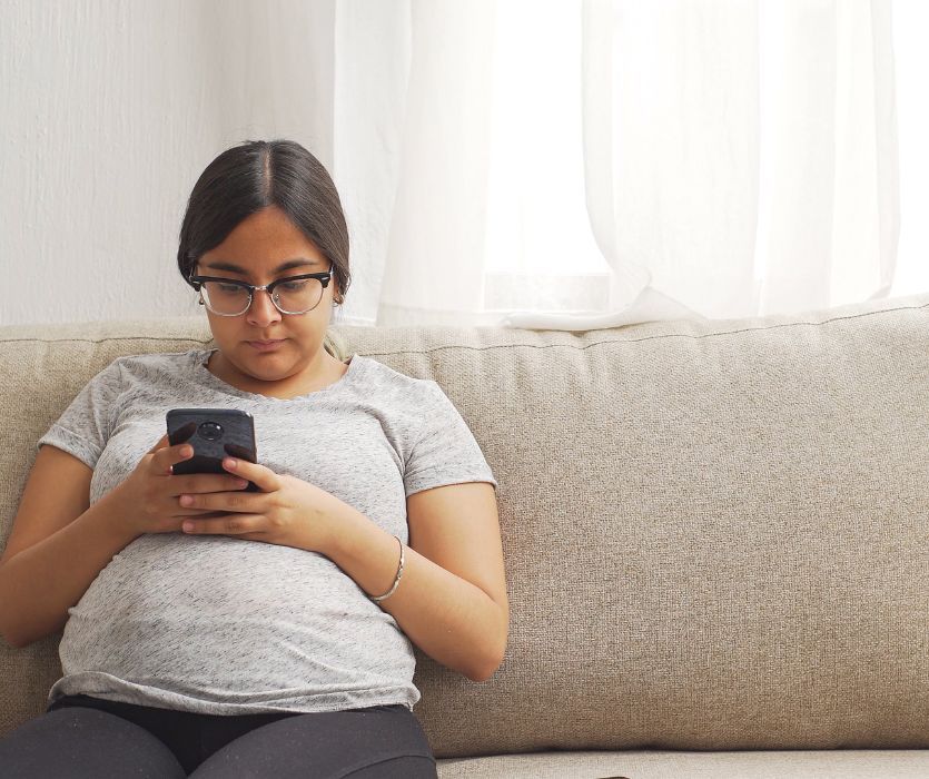 A young woman wearing glasses sits on a couch while looking down at a smartphone in her hands.