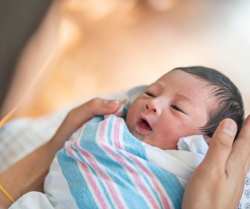 A newborn baby swaddled in a pink and blue striped hospital blanket, being held by an adult.