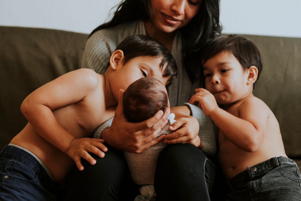 A mother cradles a newborn while her two young sons lean in to look at their new sibling.