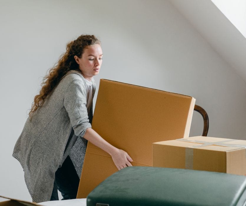 A pregnant woman lifting a large cardboard box while moving.