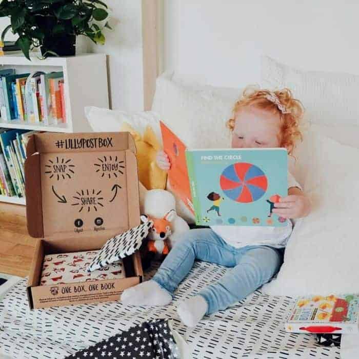 Toddler with red curly hair reading a board book next to an open Lillypost subscription box on a bed.