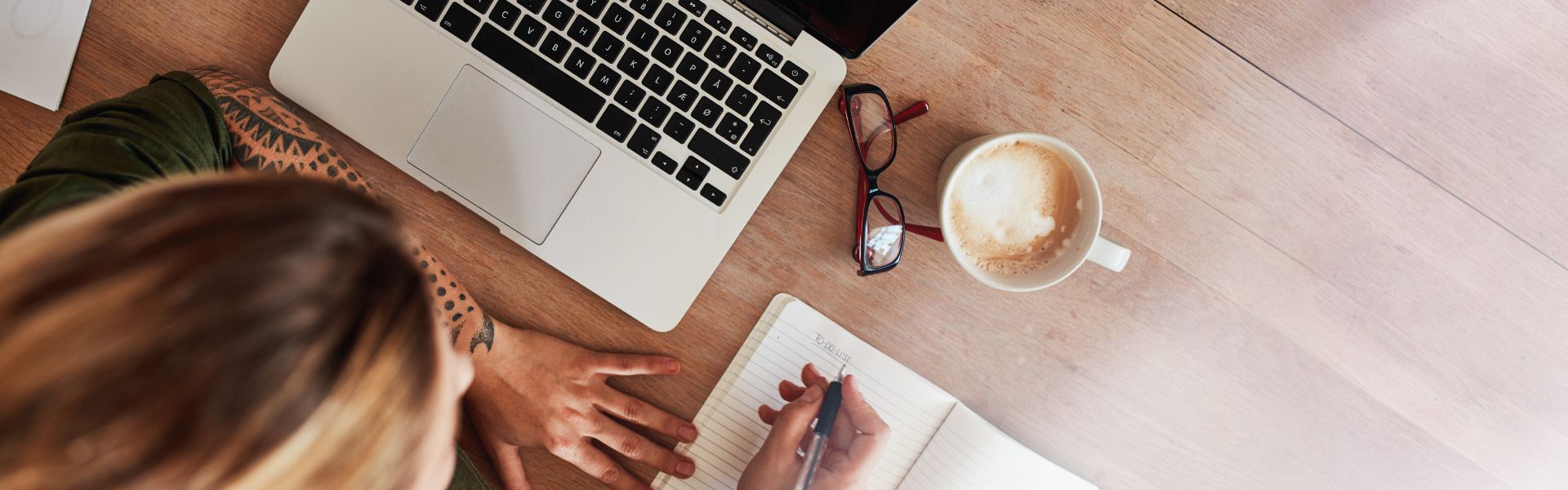 Top-down view of a person writing a to-do list in a notebook next to a laptop, eyeglasses, and a cup of coffee.