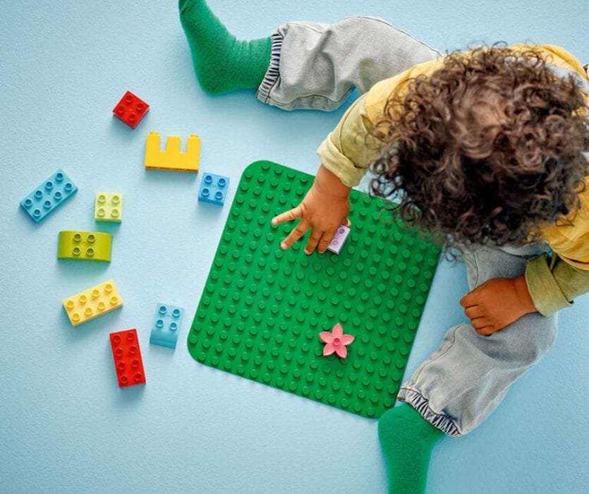 Child with curly hair plays with colorful building blocks and a green baseplate on a light blue floor.