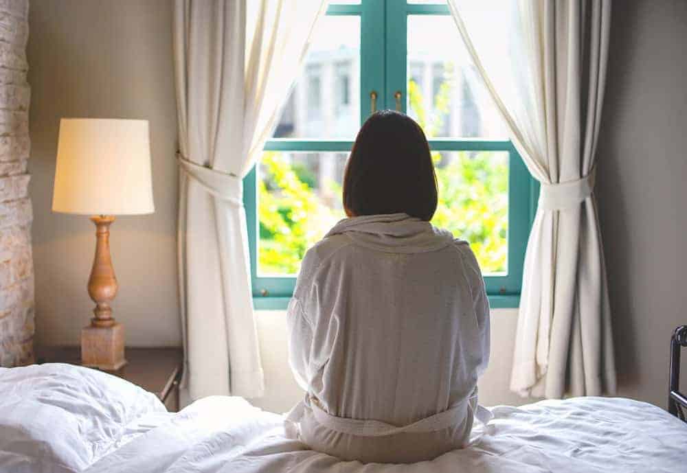 A person in a white robe sits on a bed, facing away and looking out a window with green frames.