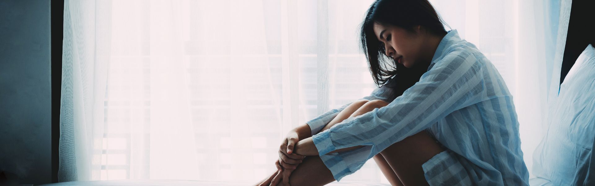 A woman sits on a bed with her head bowed and arms wrapped around her knees in front of a bright window.