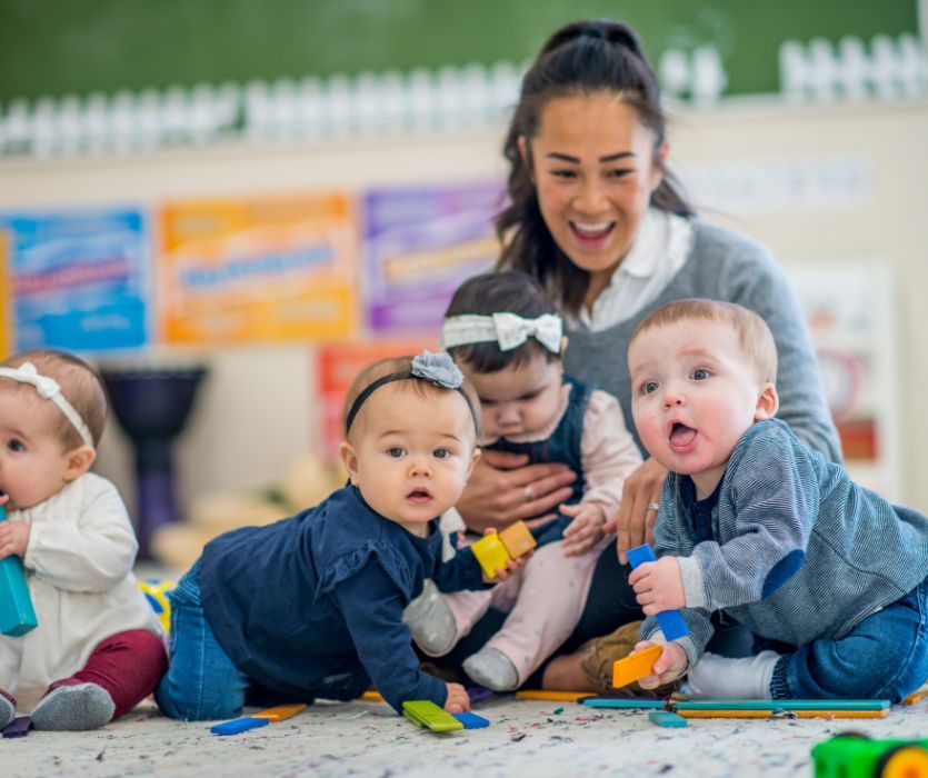 A smiling daycare teacher watches four infants playing with colorful wooden building blocks on a rug.