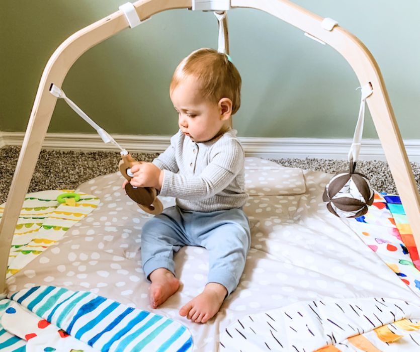 A baby sits on the Lovevery baby play gym mat, holding a hanging wooden toy while other sensory toys dangle overhead on the patterned play space.
