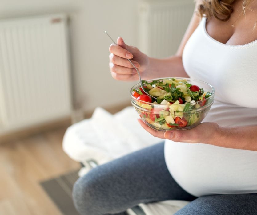 Pregnant person holding a glass bowl of fresh vegetable salad with tomatoes and greens.