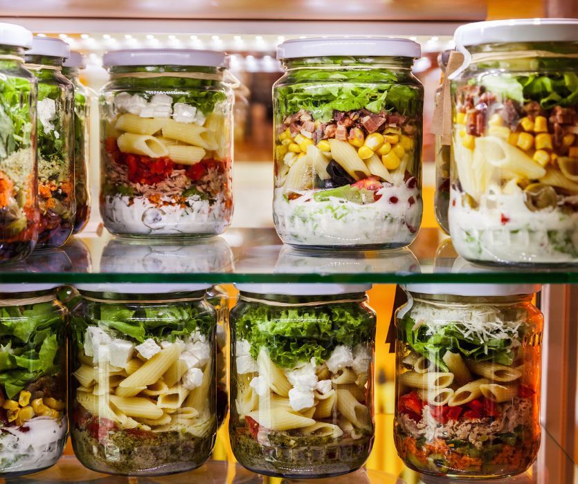 Two shelves of glass mason jars filled with layered pasta salads including lettuce, corn, penne, and dressing.