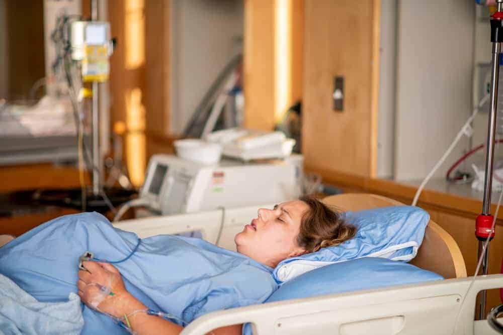 A pregnant woman in a hospital gown lies in a delivery bed with her eyes closed during labor.