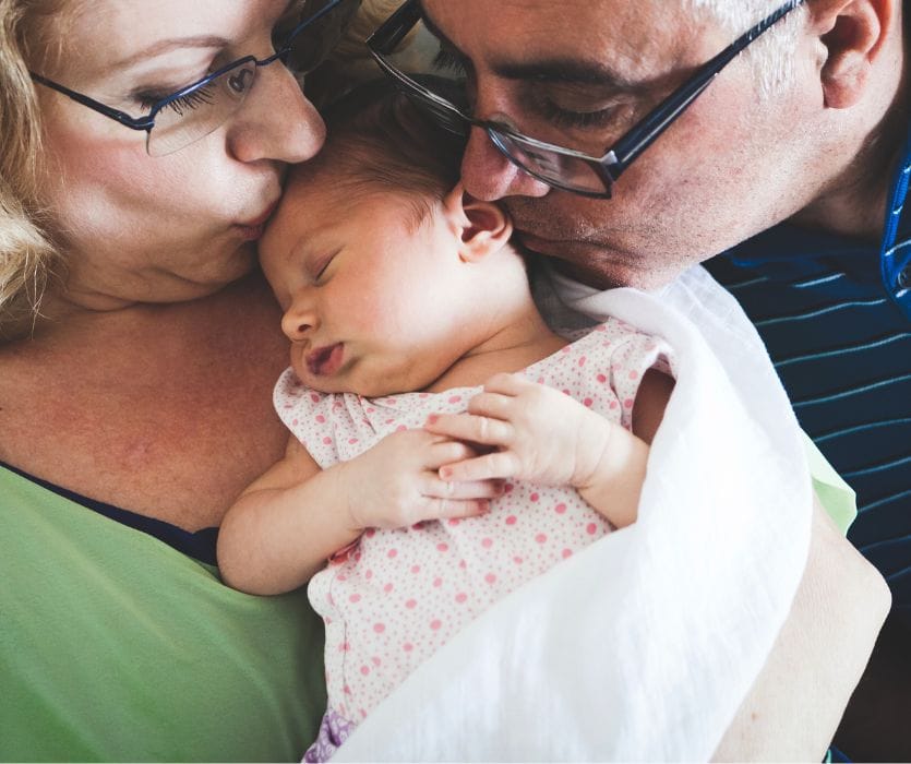 An older man and woman in glasses kiss a sleeping newborn baby's head.
