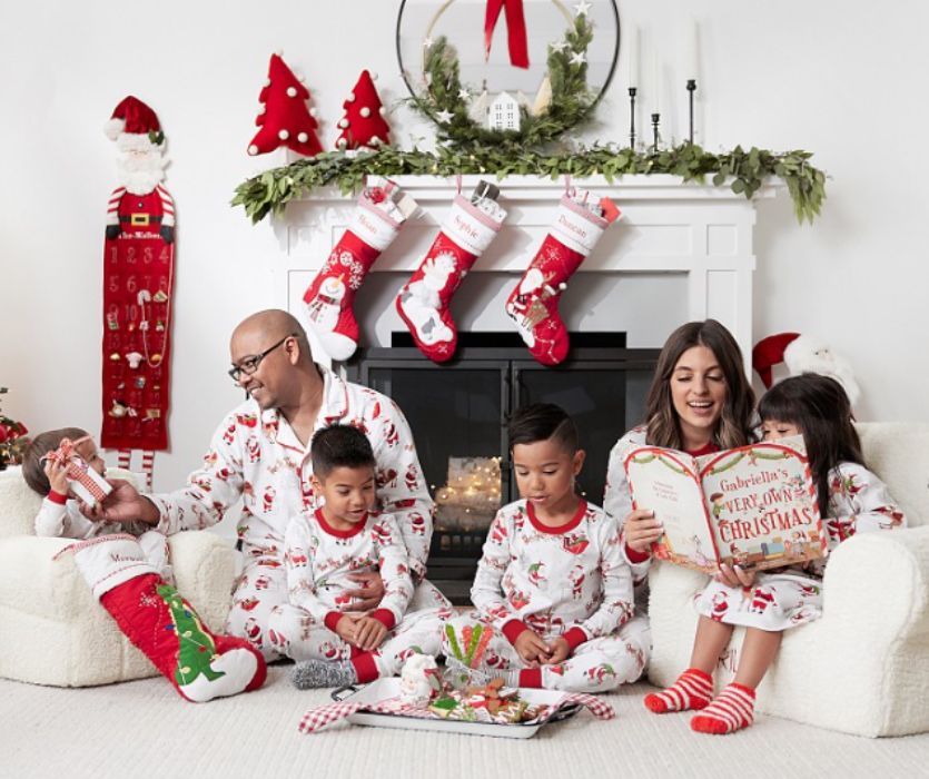A family wearing matching Santa Claus pajamas sits by a decorated fireplace reading a Christmas book.