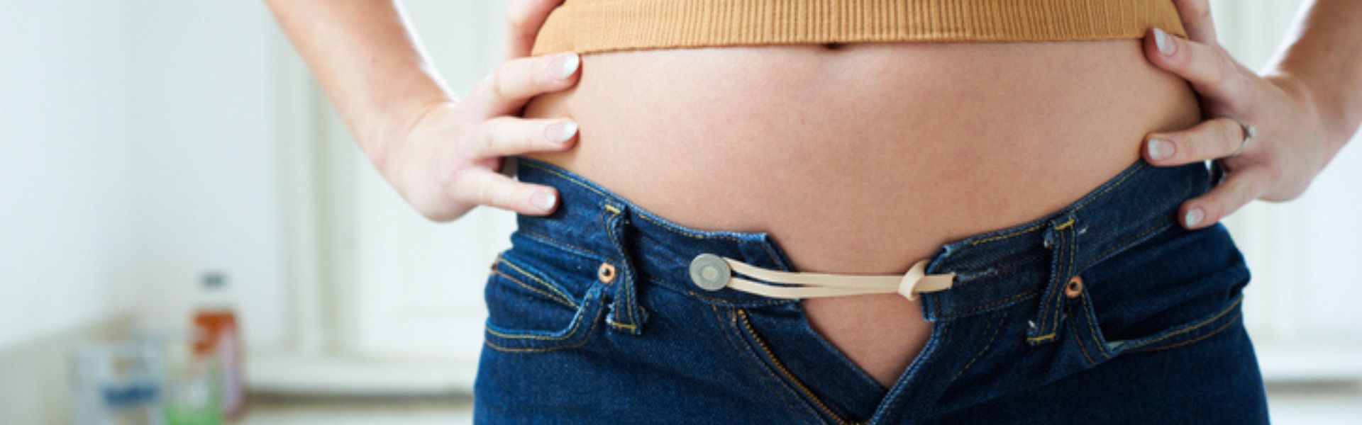 Close-up of a pregnant person using a rubber band to extend the waist of unbuttoned blue jeans.