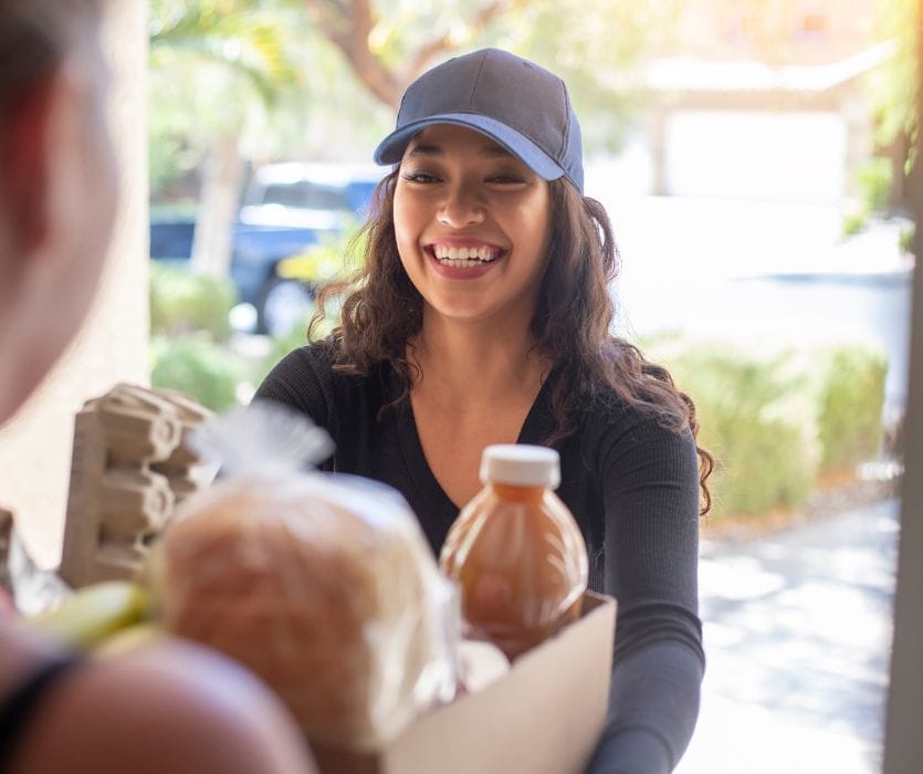 Smiling woman in a baseball cap delivering a box of groceries including bread and juice to a person's doorstep.