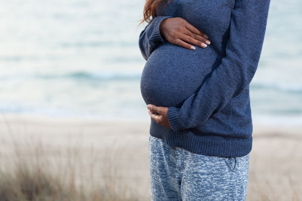 A close-up of a pregnant person standing on a beach, cradling their stomach with both hands.
