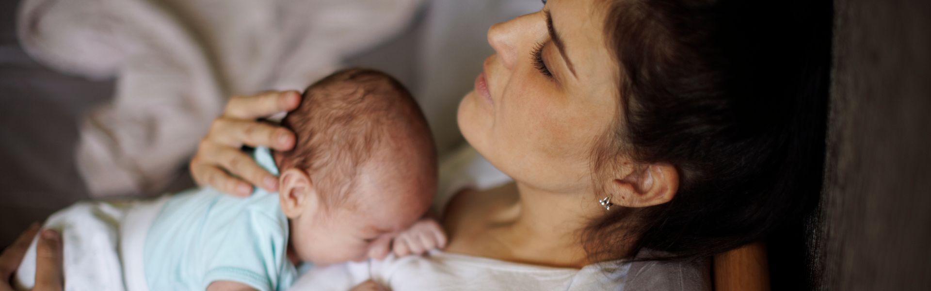 A tired-looking mother rests her eyes while holding her crying newborn baby against her chest.