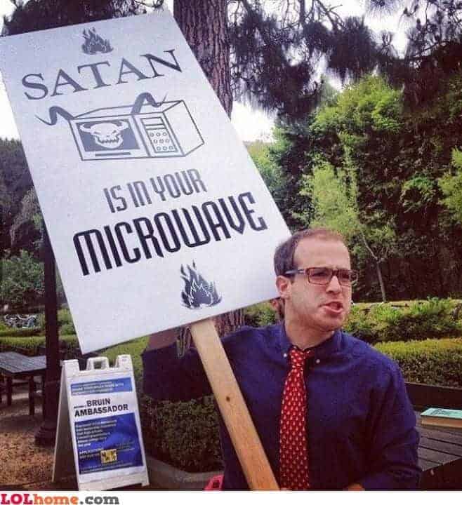 A man in a tie holds a sign that reads, Satan is in your microwave, with an illustration of a demon inside a microwave.
