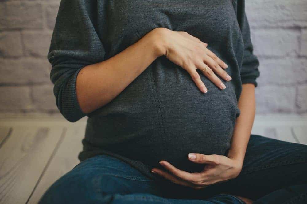 A person sitting cross-legged in a grey sweater cradle their pregnant belly with both hands.