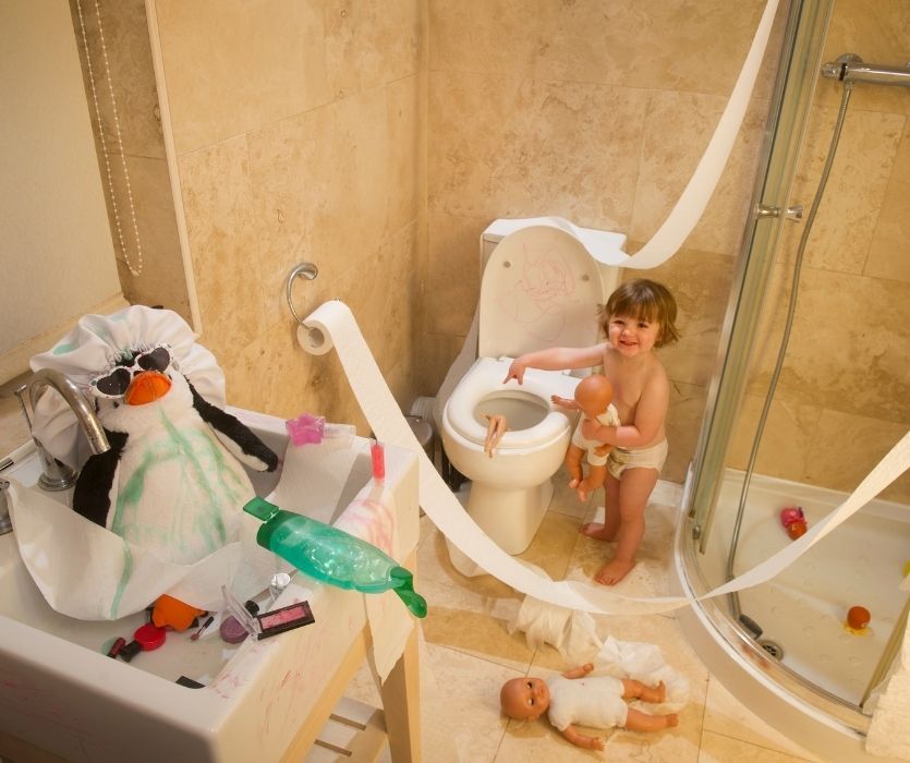 A toddler makes a mess with toilet paper, dolls, and makeup in a bathroom alongside a plush penguin in the sink.