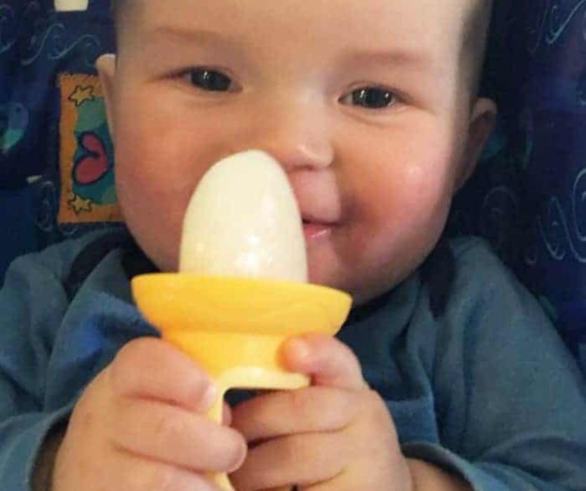 Smiling baby holding a white frozen milksicle in a yellow teether handle.