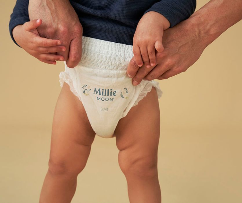 A toddler wearing white Millie Moon training pants being held at the waist by an adult.