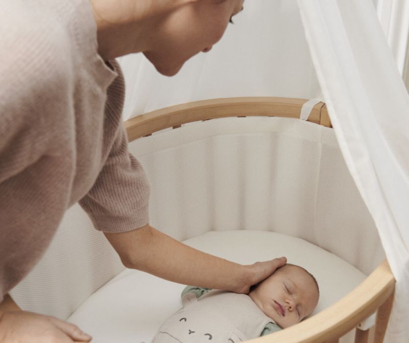 A mother gently touches the head of a sleeping baby in an oval wooden mini-crib with mesh siding and a white canopy.