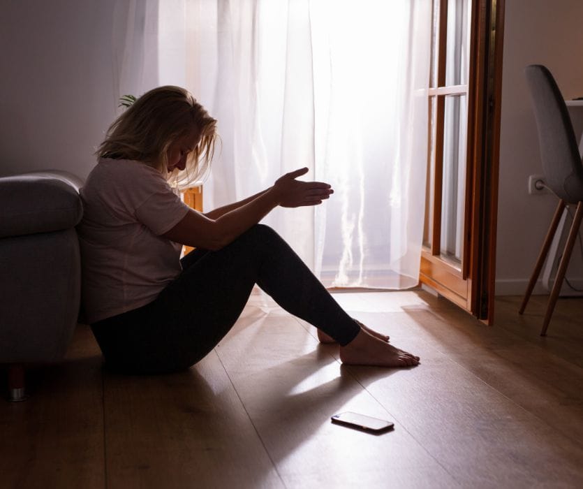 A woman sits on the floor in silhouette, head bowed and hands clasped, appearing distressed with a phone nearby.