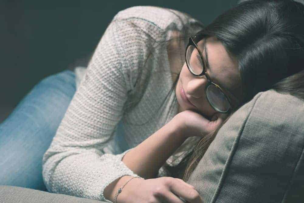 A woman with dark hair and glasses leaning her head against a sofa cushion with a somber expression.