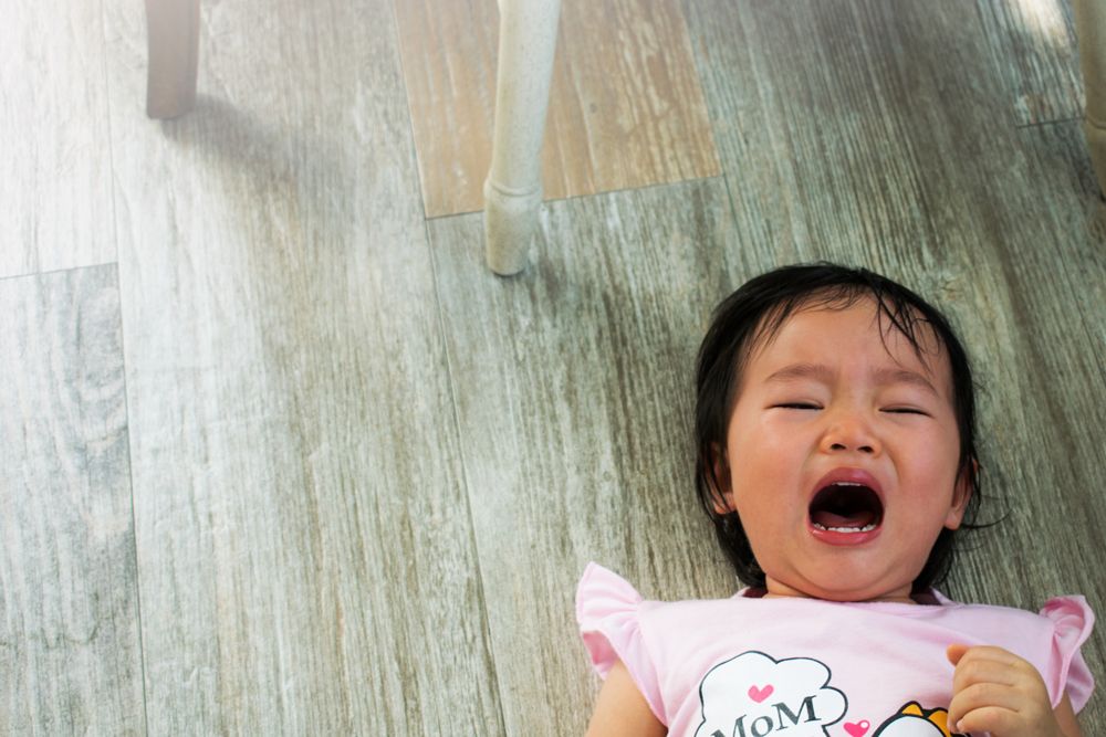 A crying toddler lying on a wooden floor, mouth open, as if having a tantrum.