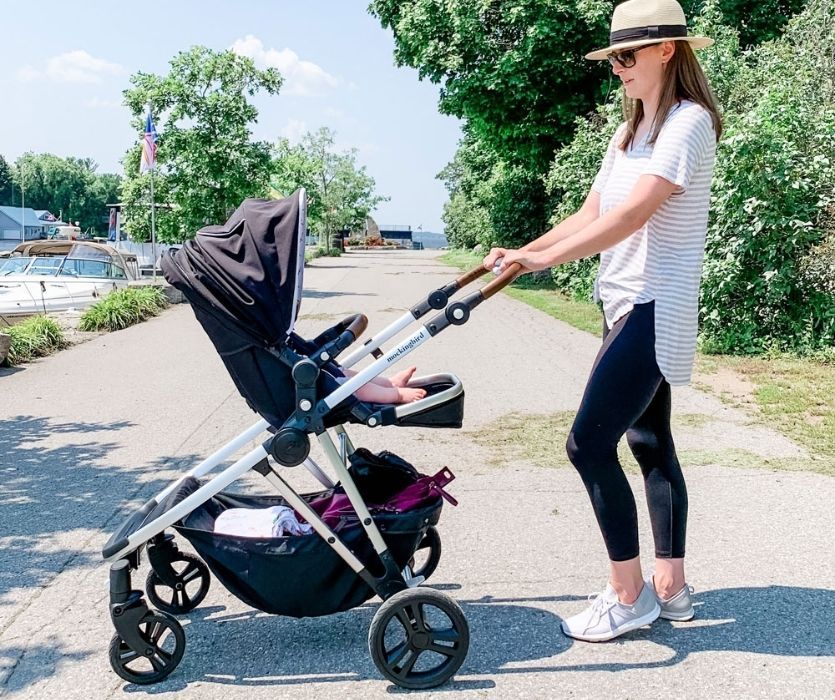 A woman in a sun hat pushes a black and silver Mockingbird stroller along a paved path near a lake harbor.