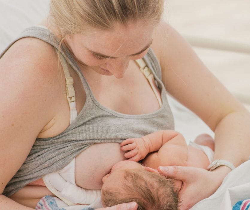 A blonde woman in a grey tank top looks down while breastfeeding her newborn infant.
