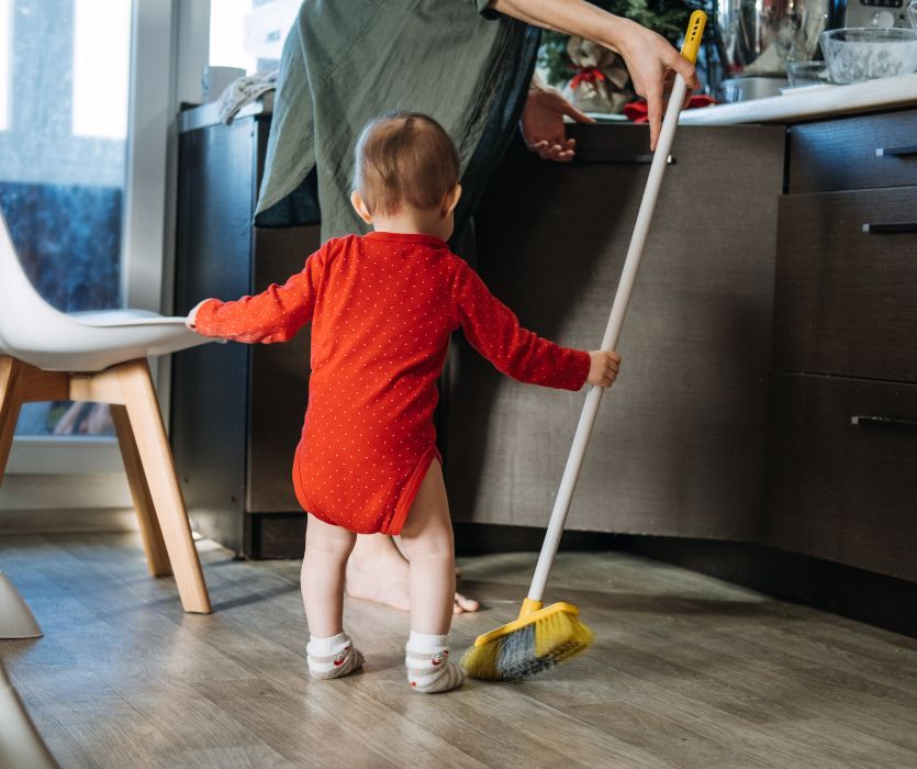 A toddler in a red onesie helps their mother sweep the kitchen floor with a yellow broom.