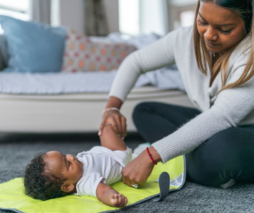 A mother sits on a carpet, changing the cloth diaper of her baby lying on a yellow mat.