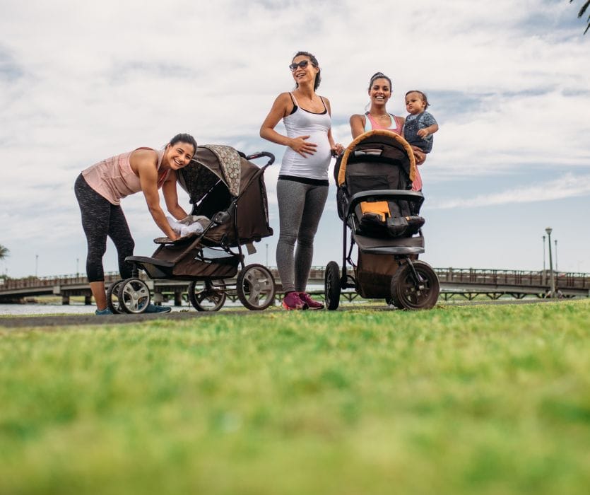 Three women posing outdoors in athletic wear with two strollers and a toddler. One woman is pregnant.