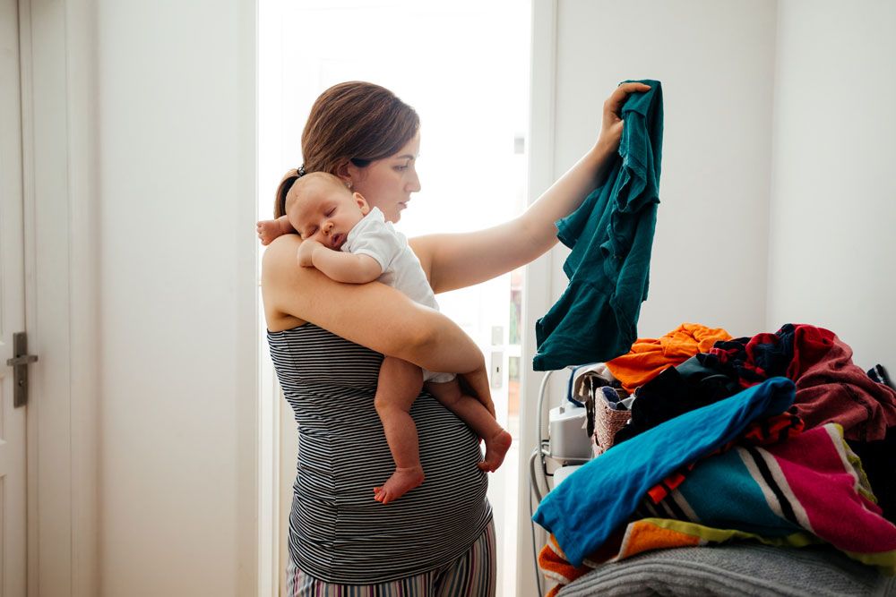 A mother carries a sleeping baby on her shoulder while sorting through a large pile of laundry.